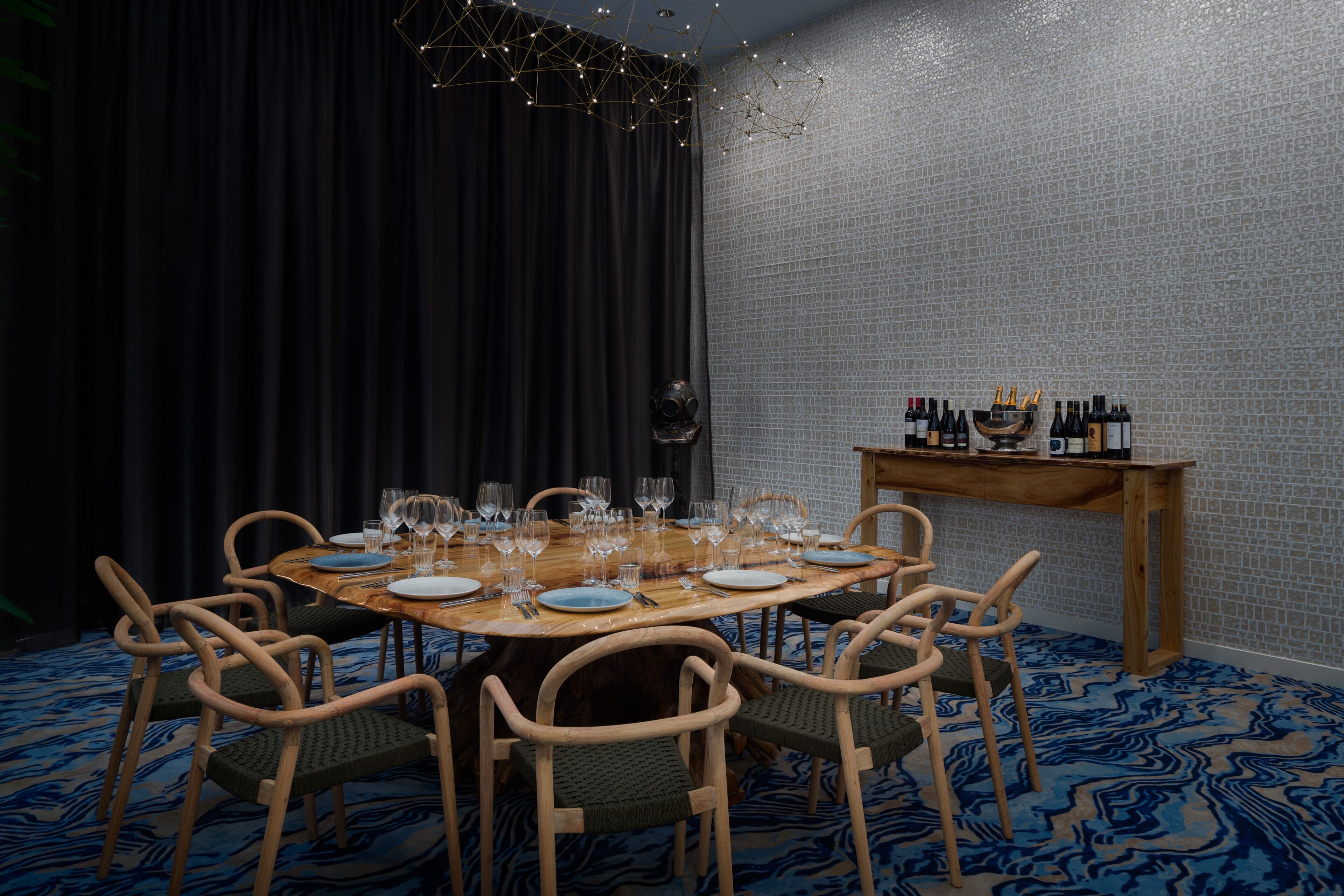 A dining area with a round table set for six, surrounded by wooden chairs. A wooden side table holds several bottles and glasses. The room features a patterned carpet and a textured wall.