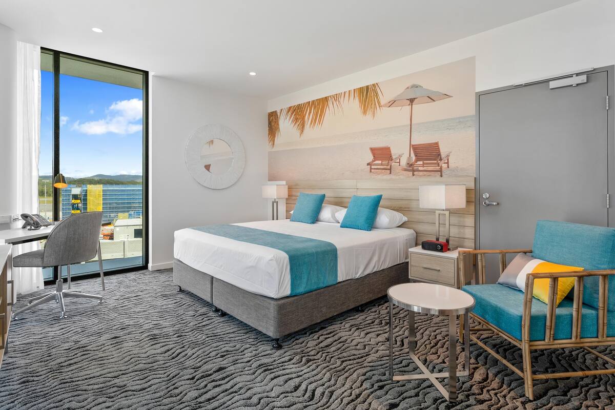 Ein modernes Hotelzimmer mit einem großen Bett, einem Schreibtisch und einem Stuhl. Durch das Fenster bietet es einen Blick auf den Strand.