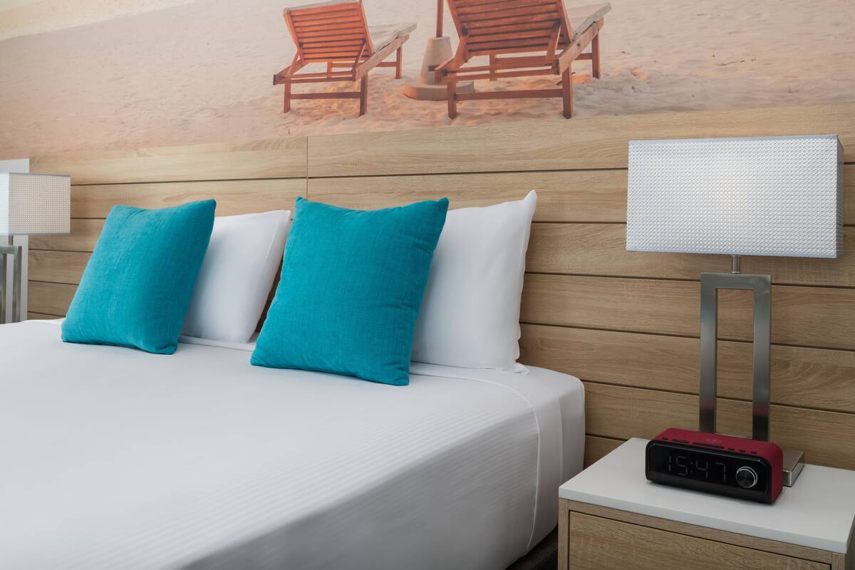 Ein Hotelzimmer mit einem Bett, zwei türkisfarbenen Kissen, einem weißen Kissen, einem Nachttisch mit rotem Wecker und einem Wandbild von Liegestühlen.
