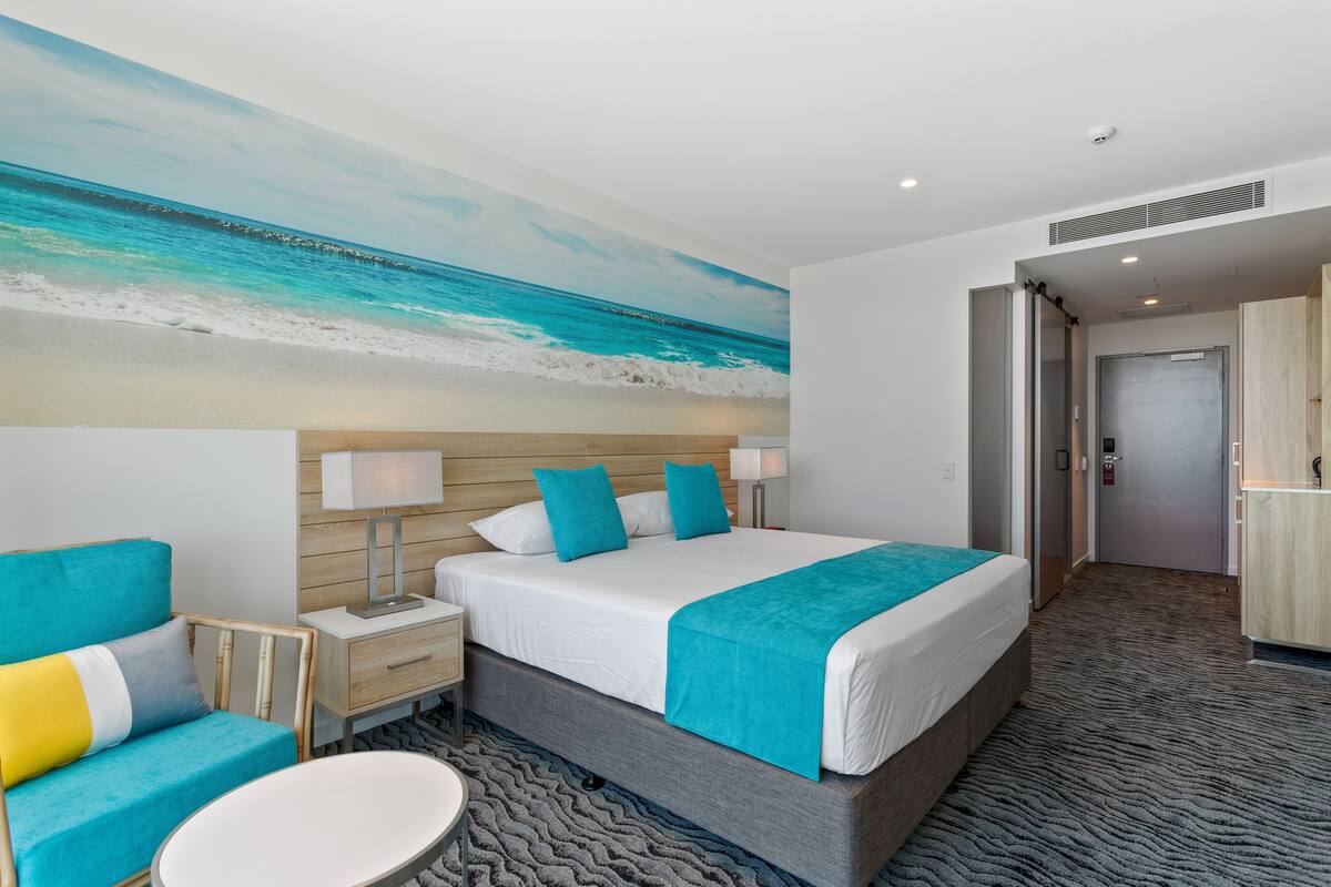 Ein Hotelzimmer mit Bett, Nachttisch, Sessel, rundem Tisch und einem Bild mit Strandmotiv.