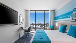 A hotel room with a large bed, a desk, a chair, a TV, and a view of the city through large windows.