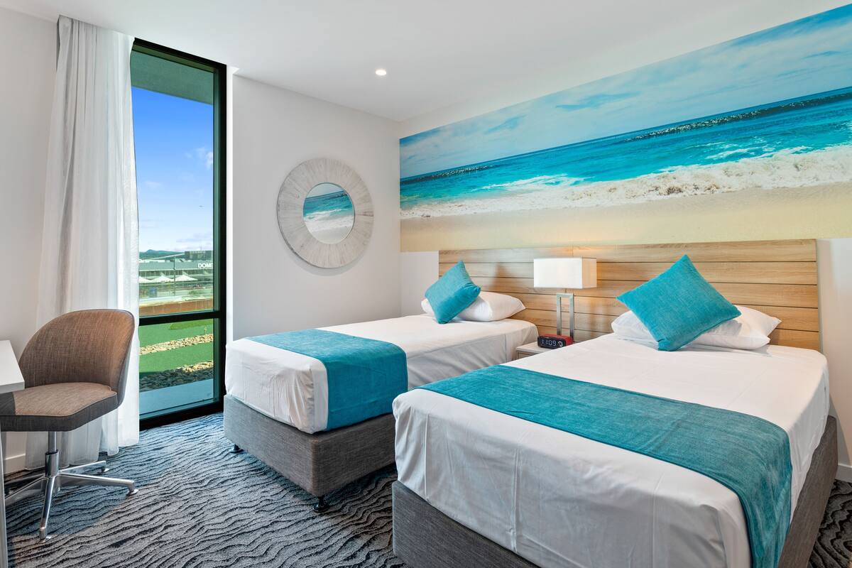 Ein Hotelzimmer mit zwei Betten, einem Schreibtisch und einem großen Fenster mit Blick auf den Strand.