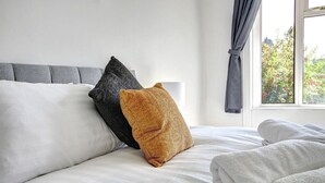 Una cama bien hecha con sábanas blancas, un cabecero gris y dos almohadas, una gris y otra mostaza.