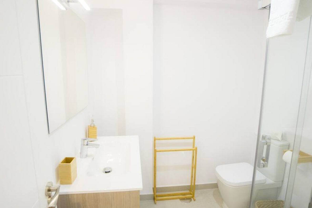 Ein modernes Badezimmer mit einem weißen Waschbecken, Spiegel und Toilette. Neben dem Waschbecken befindet sich ein Holzregal und eine Dusche mit Glaswand.