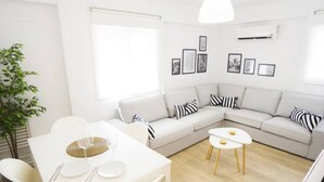 Ett modernt vardagsrum med en vit soffa, ett matbord med stolar och ett litet soffbord. På väggen hänger inramade tavlor och i hörnet står en växt.
