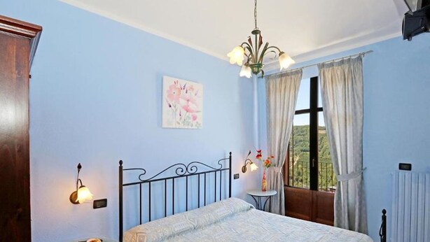 Een slaapkamer met een bed, een klein tafeltje, een nachtkastje, een raam met gordijnen, een schilderij aan de muur en een radiator.