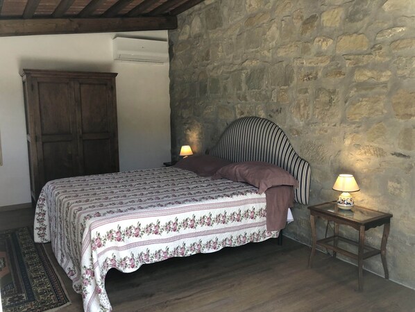 A bedroom with a bed, two bedside lamps, a wooden nightstand, a rug, a stone wall, and a wooden door.