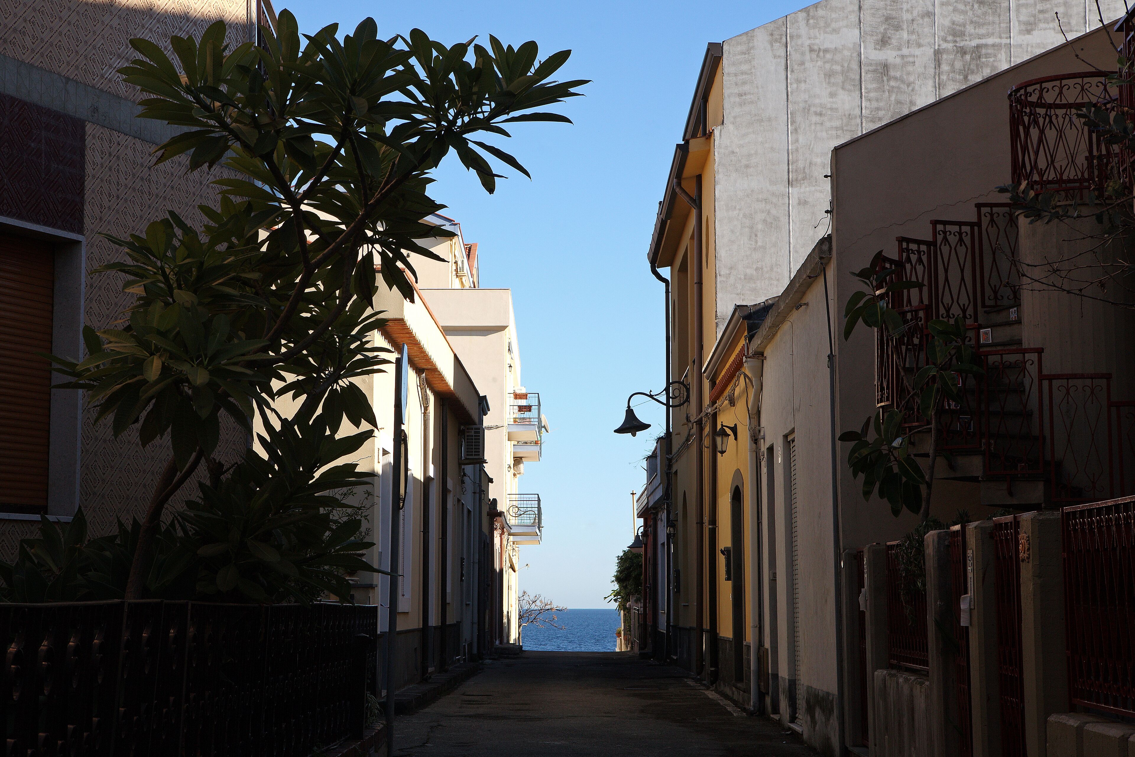 Eine enge Straße mit Häusern an beiden Seiten, ein klarer Himmel und ein Blick auf das Meer in der Ferne.