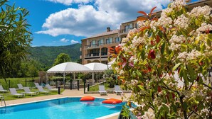 Piscina com espreguiçadeiras e guarda-sóis, rodeada por vegetação exuberante e um edifício com varandas.