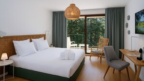Quarto de hotel com cama, escrivaninha, cadeira e varanda com vista para árvores.