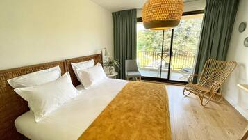 A hotel room with a large bed, a wooden headboard, a hanging light fixture, a balcony with a chair, and a view of greenery outside.