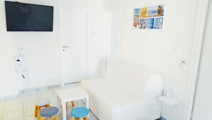 Een moderne, witte woonkamer met een grote bank, een klein tafeltje, twee krukken, een flatscreen televisie aan de muur en een kunstwerk.