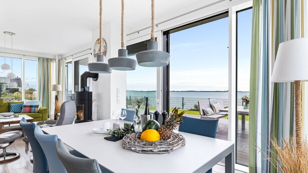 A modern dining area with a white table, blue chairs, and a fruit centerpiece. - Villa Seemööv - waterfront, terrace, whirlpool, sauna, fireplace, e-charging (Olpenitz)
