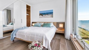 A bedroom with a large bed, a wooden wardrobe, a bedside table with a lamp, and a view of the sea through a large window. - Villa Seemööv - waterfront, terrace, whirlpool, sauna, fireplace, e-charging (Olpenitz)