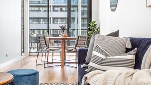 Una sala de estar moderna con un sofá azul oscuro, una mesa redonda de madera con sillas, y un balcón con vista a un edificio.