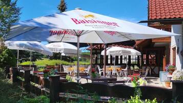 Un espace extérieur aménagé avec des tables et des chaises sous de grands parasols, un bâtiment avec un toit recouvert de tuiles rouges et une végétation luxuriante au premier plan.