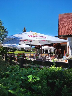 An outdoor seating area with tables and chairs under large umbrellas, a building with a red-tiled roof, and lush greenery in the foreground. - Einzelzimmer - Stahlecker Hof (Lichtenstein)
