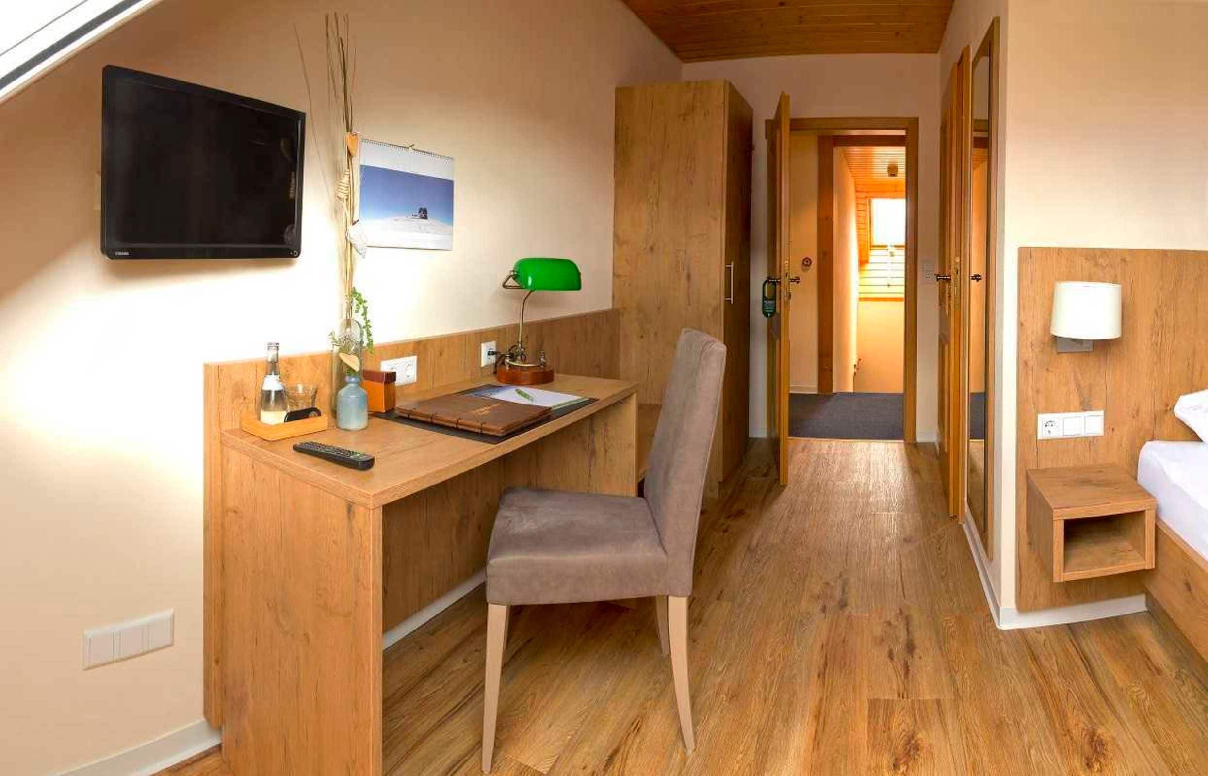 Une pièce de taille modeste, comprenant un bureau en bois, une chaise, un lit et un téléviseur fixé au mur.