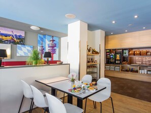 A modern cafe with a counter, seating area, and a display of pastries and beverages. - B&B Hotel München City-Nord (Munich)