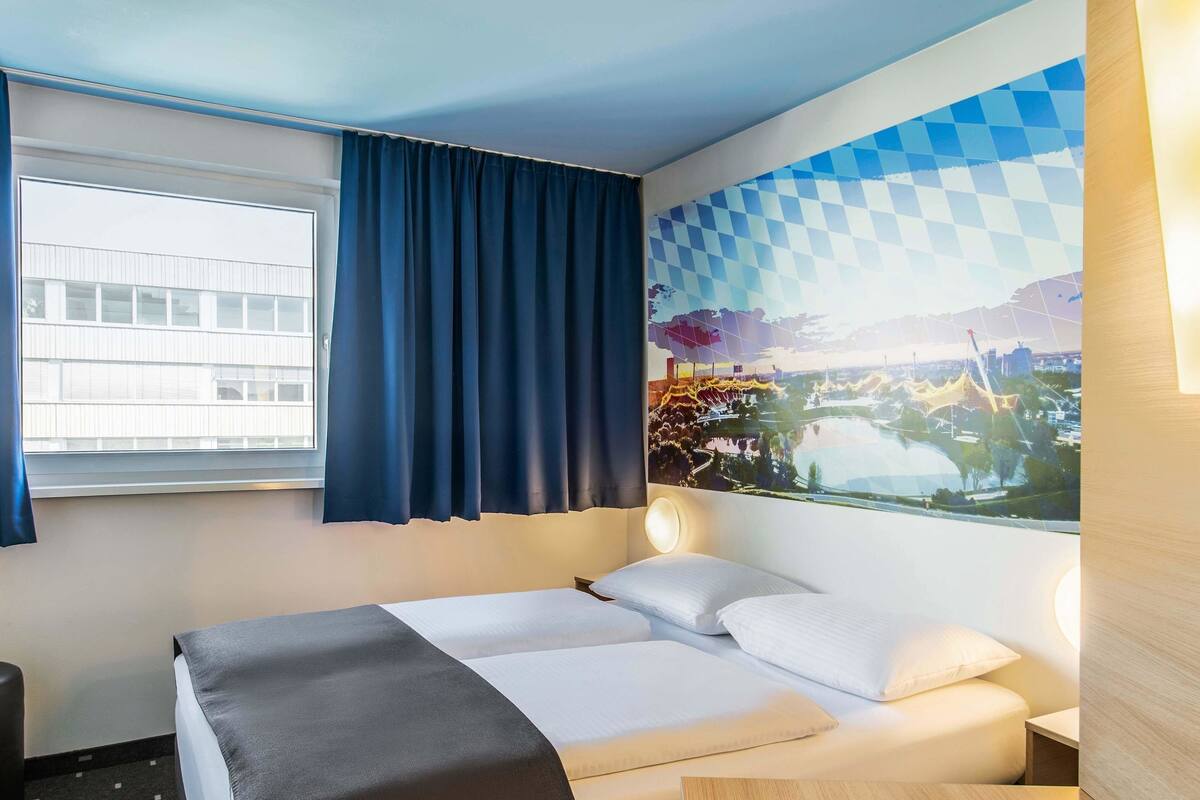 A hotel room with a bed, a desk, a window with curtains, and a wall with a cityscape mural.
