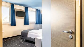 Una habitación de hotel con cama, una mesita de noche pequeña, un televisor montado en la pared y una ventana con cortinas.