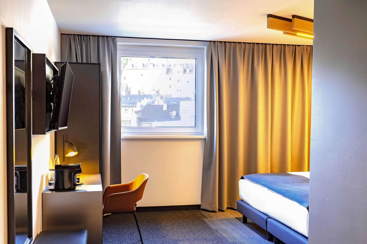 A hotel room with a bed, a desk with a lamp, a chair, and a TV.