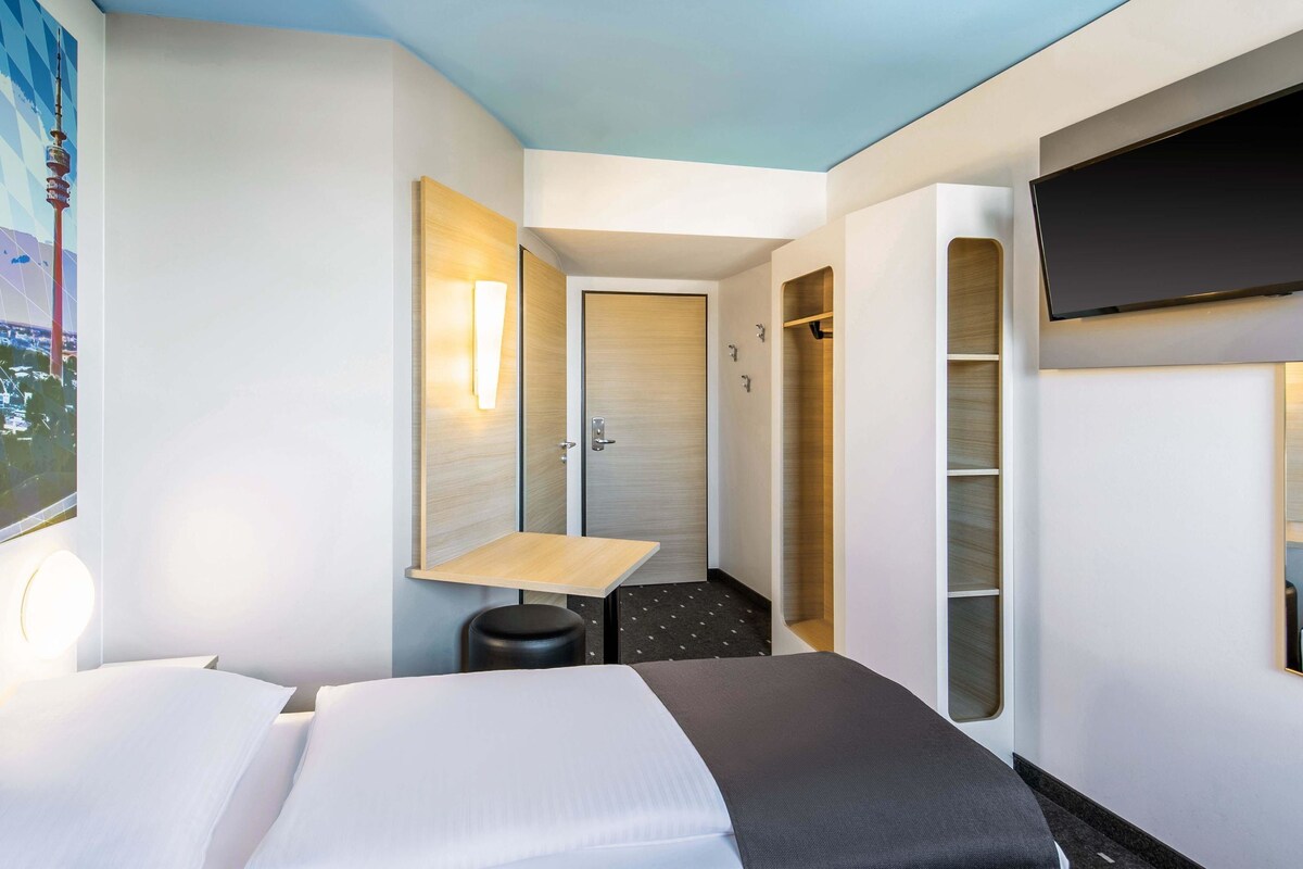 A hotel room with a bed, a desk, a chair, a TV mounted on the wall, and a door leading to a closet.