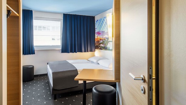 Habitación de hotel pequeña con cama, un escritorio, una silla, ventana con cortinas y vista a un edificio.