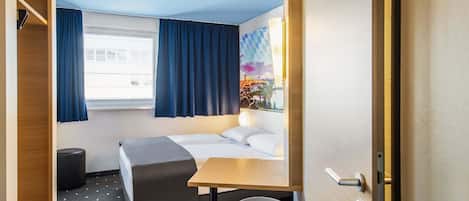 A compact hotel room with a bed, a small desk, a chair, a window with curtains, and a view of a building outside.
