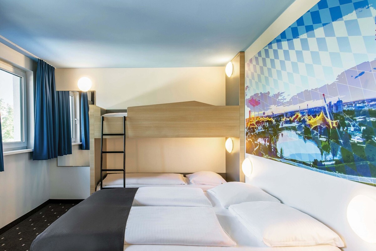 A hotel room with a bunk bed, a desk, a window with curtains, and a wall-mounted artwork.