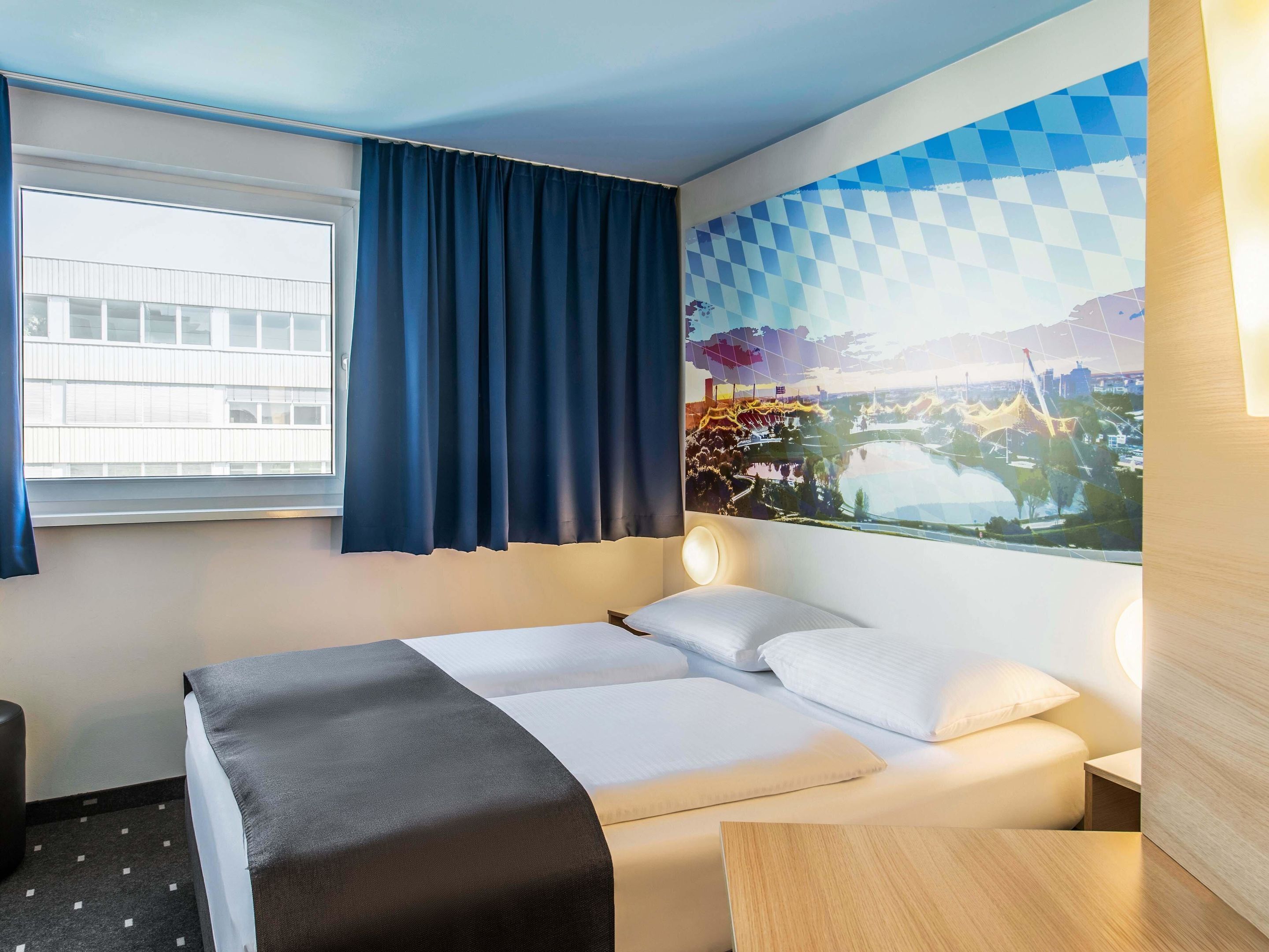 A hotel room with a bed, a desk, a window with curtains, and a wall with a cityscape mural.