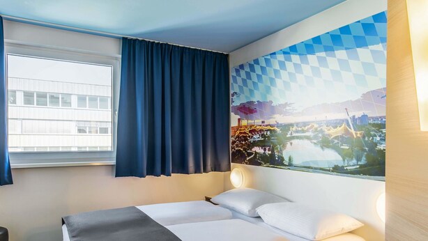 Habitación de hotel con cama, escritorio, ventana con cortinas y un mural urbano en la pared.