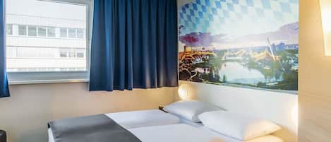 A hotel room with a bed, a desk, a window with curtains, and a wall with a cityscape mural.