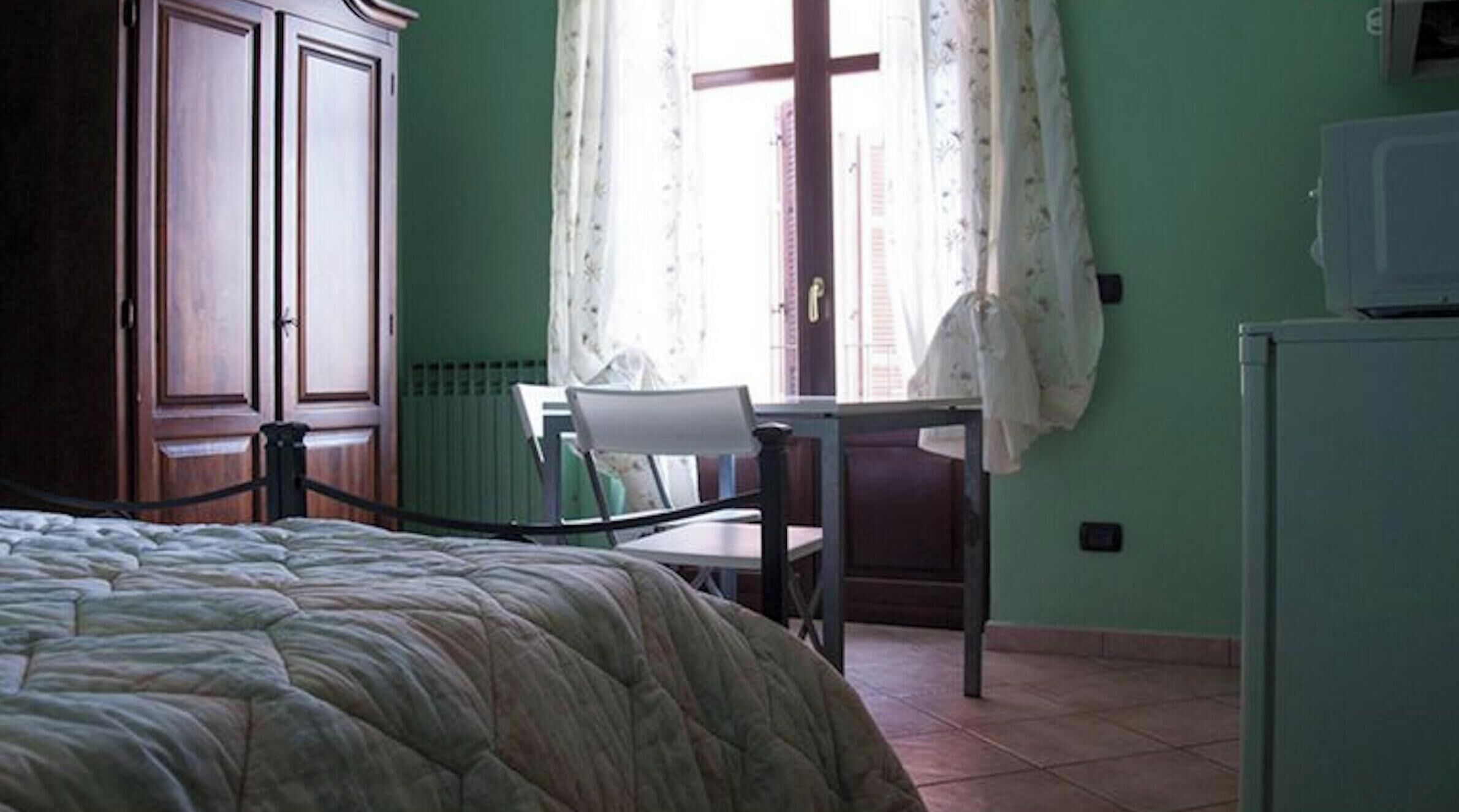 Un dormitorio con cama, una silla, un escritorio, un radiador y una ventana con cortinas.