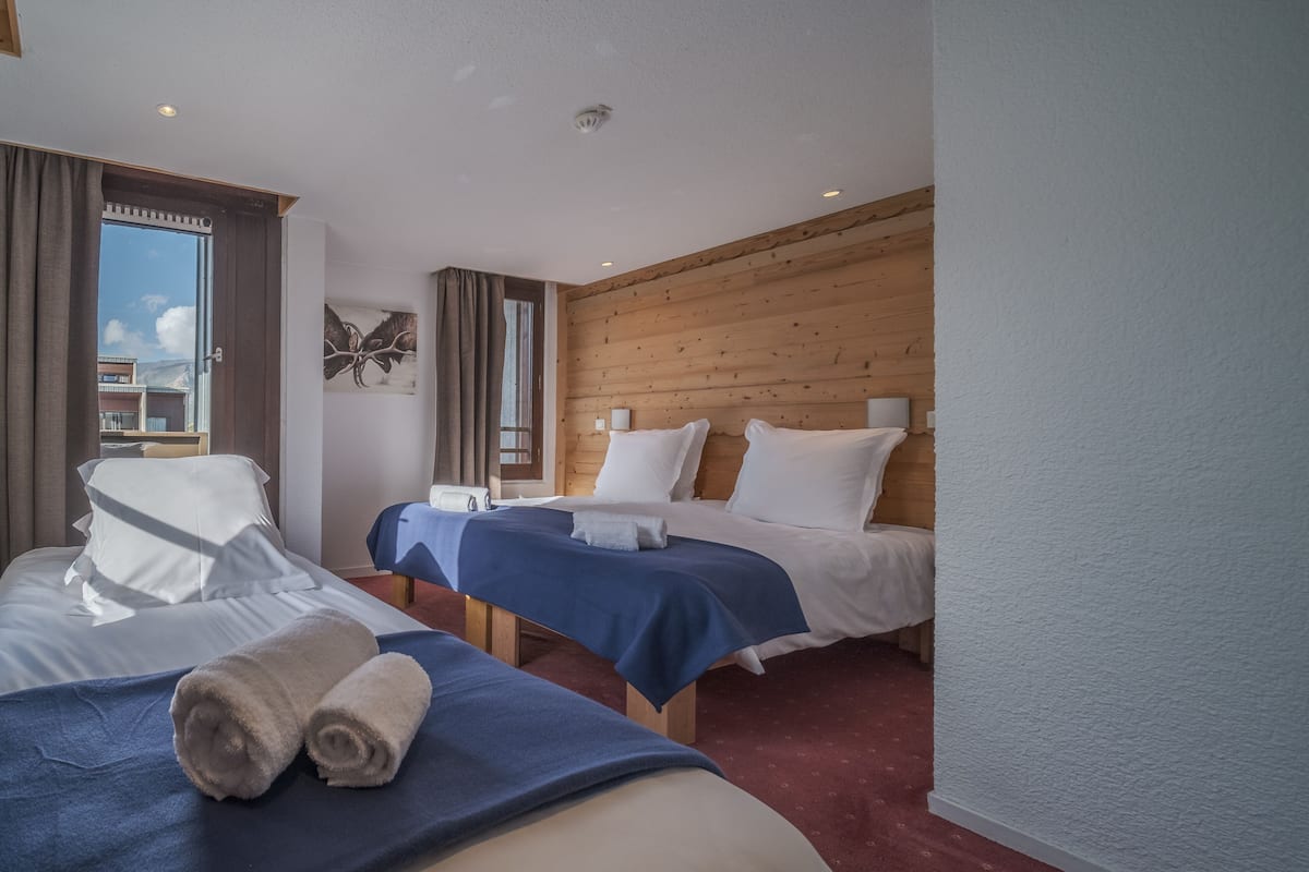 A hotel room with two beds, a large window, wooden paneling, and a view of buildings outside.
