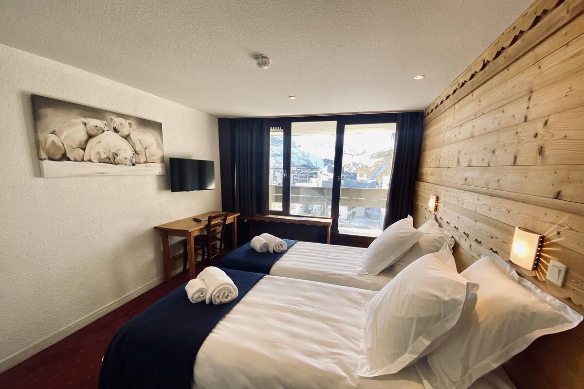 A hotel room with two beds, a desk, a chair, a TV, and a window with a view of buildings.