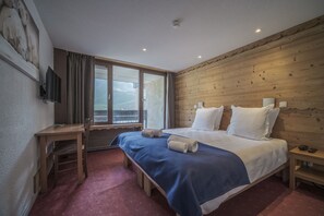 A hotel room with a wooden wall, a large bed with white pillows, a desk with a chair, and a television on the wall. - Curling Hôtel & Bar (Tignes)