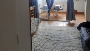Un dormitorio con una cama, una mesita y una silla. Hay una ventana con cortinas, y una puerta que da a otra habitación.