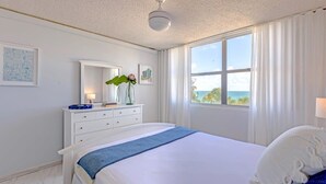 Une chambre à coucher avec un lit, une commode, un miroir, une fenêtre avec des rideaux et une vue sur la mer.