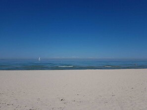 A beach with a clear sky, a sailboat in the distance, and a sandy shore.