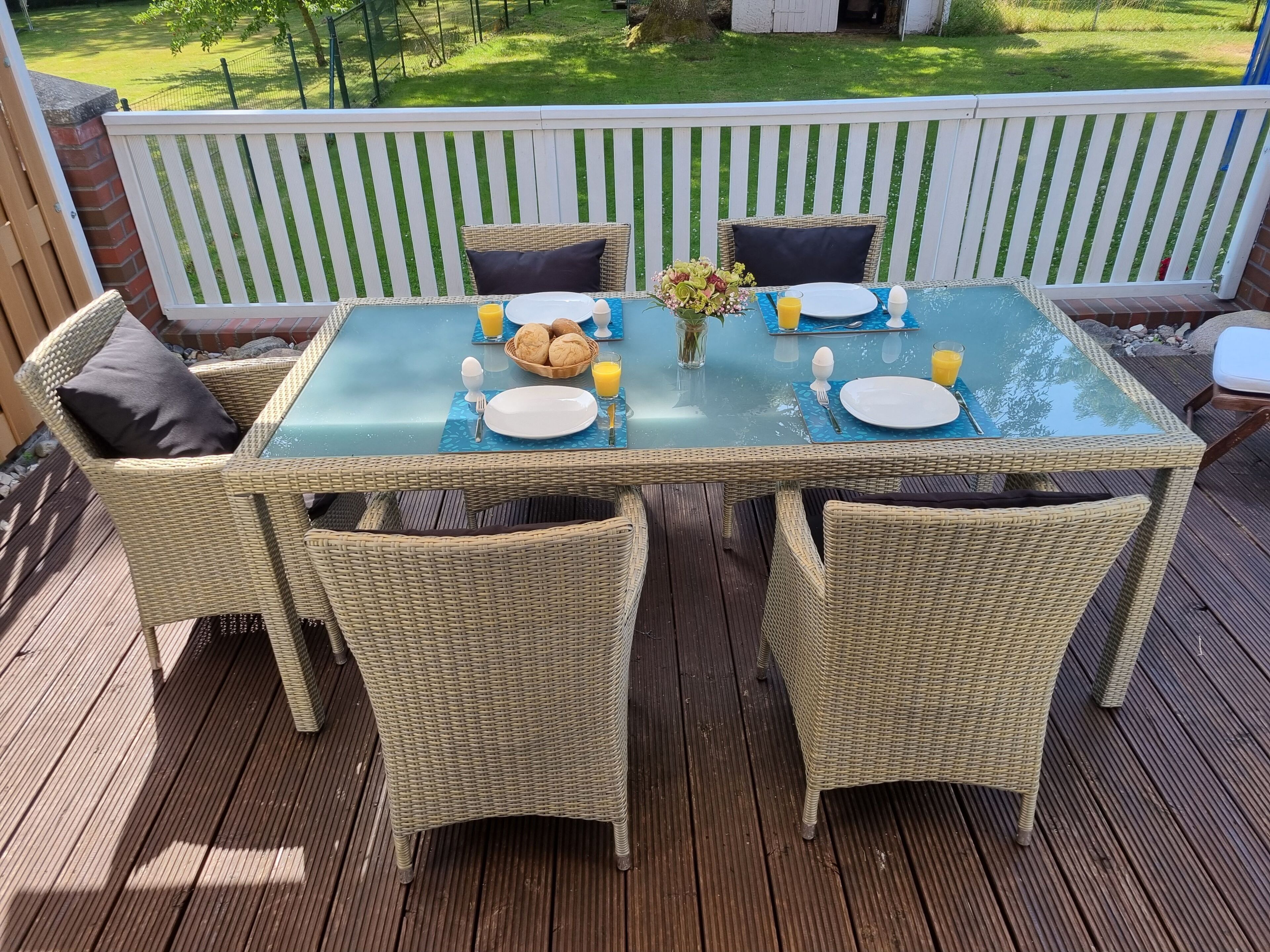 Ein Esstisch mit Geschirr, Besteck und einer Obstschale auf einer Terrasse mit Korbstühlen.