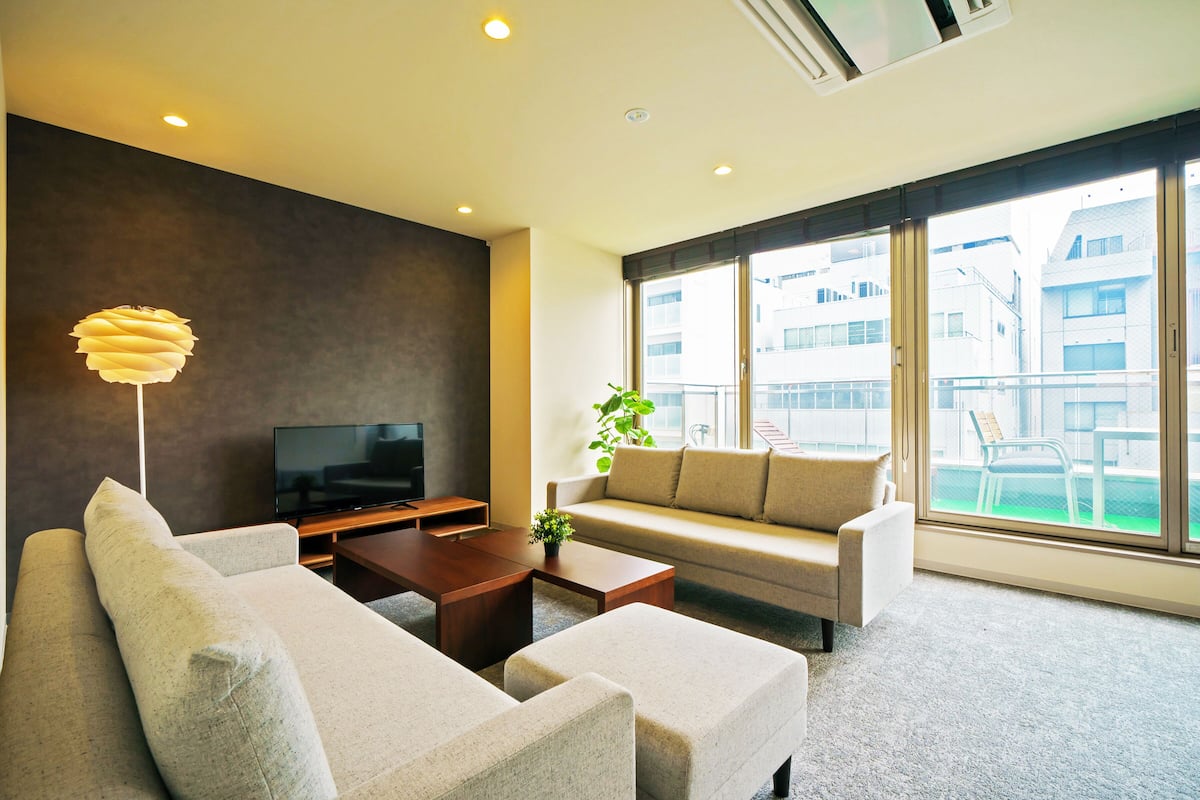 A modern living room with a sofa, two armchairs, a coffee table, a TV, and a large window with a view of buildings.