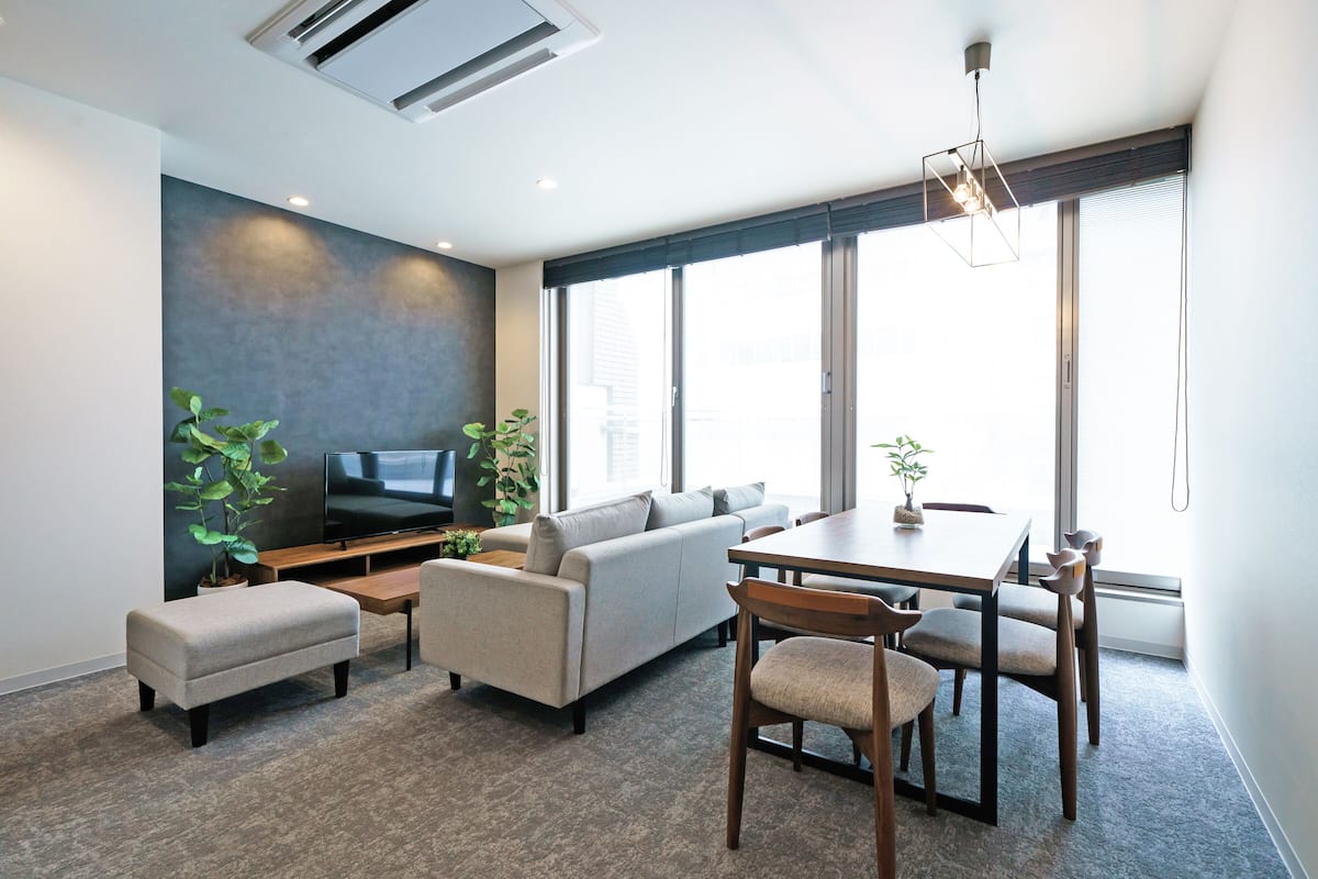 A modern living room with a sofa, ottoman, dining table, chairs, and a flat-screen TV.
