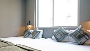 Una habitación de hotel moderna con un ventanal grande, un sofá con cojines grises y blancos, y una mesita de noche con una lámpara.