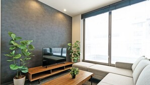 Una sala de estar moderna con un televisor de pantalla plana sobre un mueble de madera, un ventanal con vistas a edificios y dos plantas en macetas.