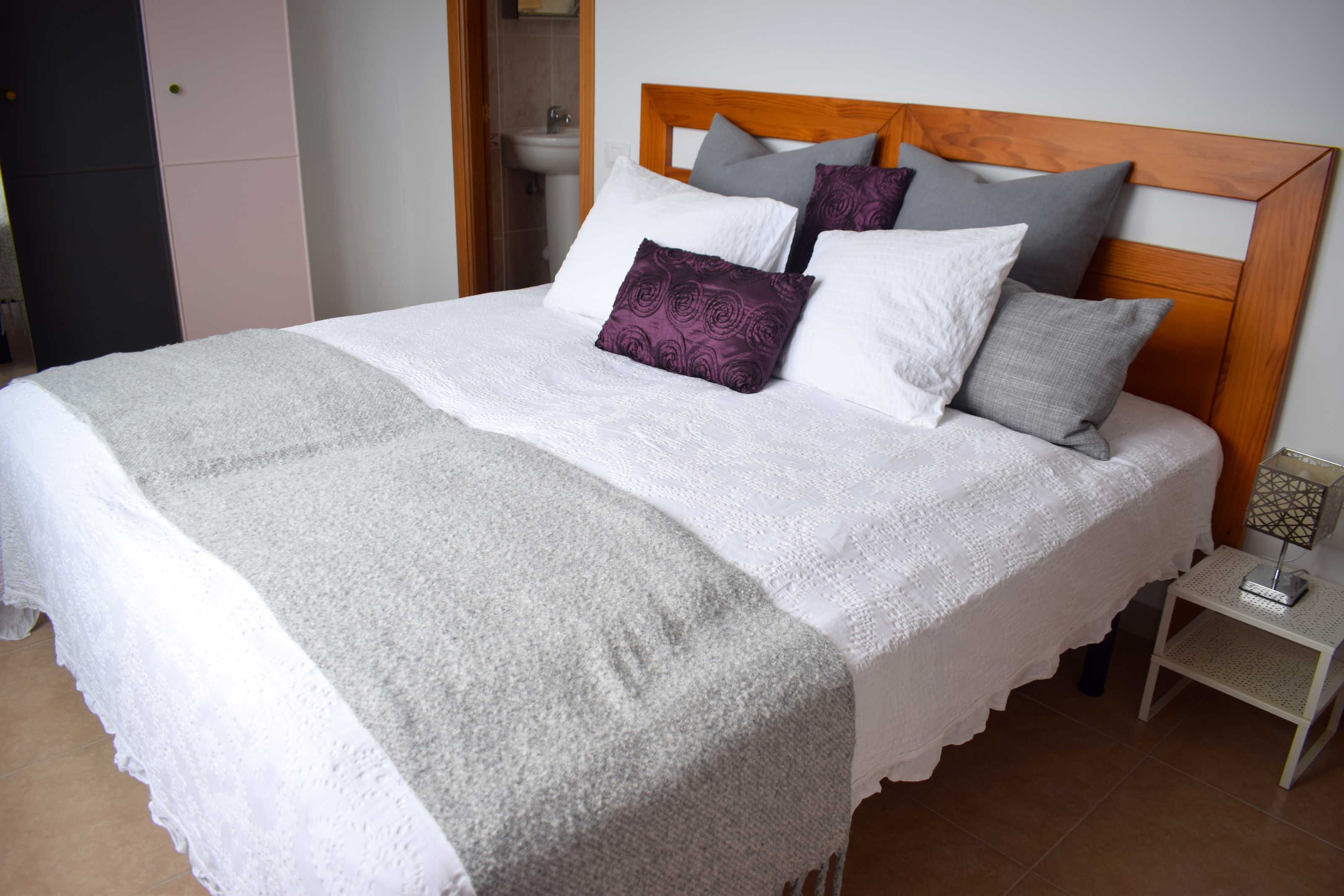Cama bem arrumada com manta branca e cinzenta, cabeceira de madeira e vários travesseiros em branco, cinza e roxo.