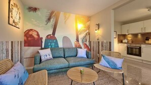 A living room with a sofa, two armchairs, a coffee table, a wall clock, and a kitchen area. - Malibu SA 57a - Malibu, 3 rooms, 65m², terrace, sauna (Scharbeutz)