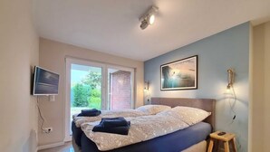 A bedroom with a large bed, a television mounted on the wall, and a window with a view of greenery. - Malibu SA 57a - Malibu, 3 rooms, 65m², terrace, sauna (Scharbeutz)