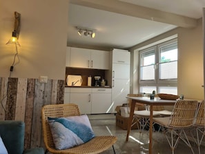Cocina compacta con armarios blancos, zona de comedor con sillas de mimbre y una ventana con persianas.