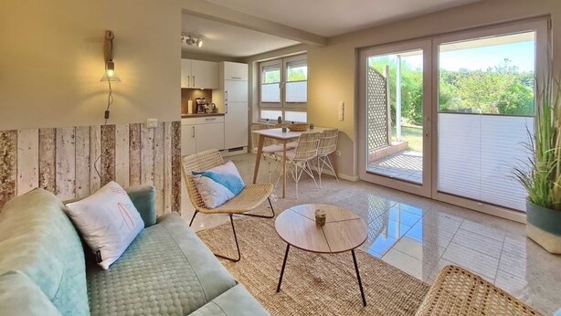 A spacious living area with a sofa, a small round table, and a dining table with chairs. There is a kitchen in the background and a sliding glass door leading to a balcony with a view of greenery. - Malibu SA 57a - Malibu, 3 rooms, 65m², terrace, sauna (Scharbeutz)